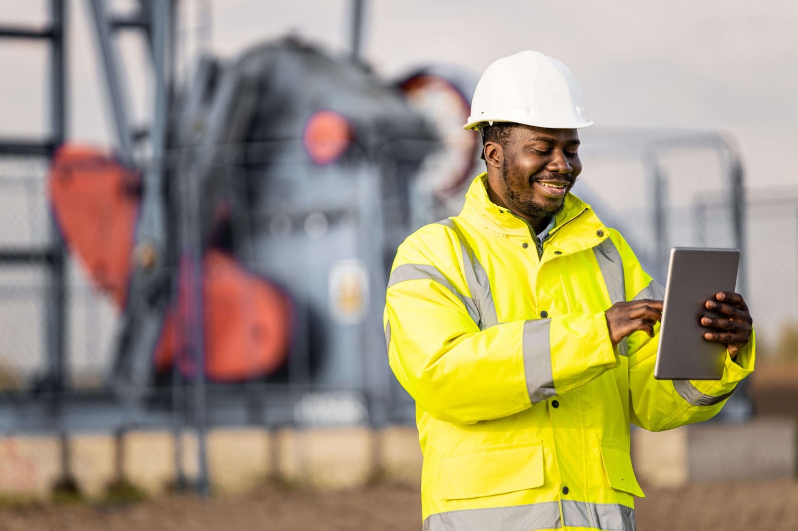 An oil industry engineer in a high-visibility jacket and hard hat using a tablet for digital monitoring and data analysis at an oil extraction site.